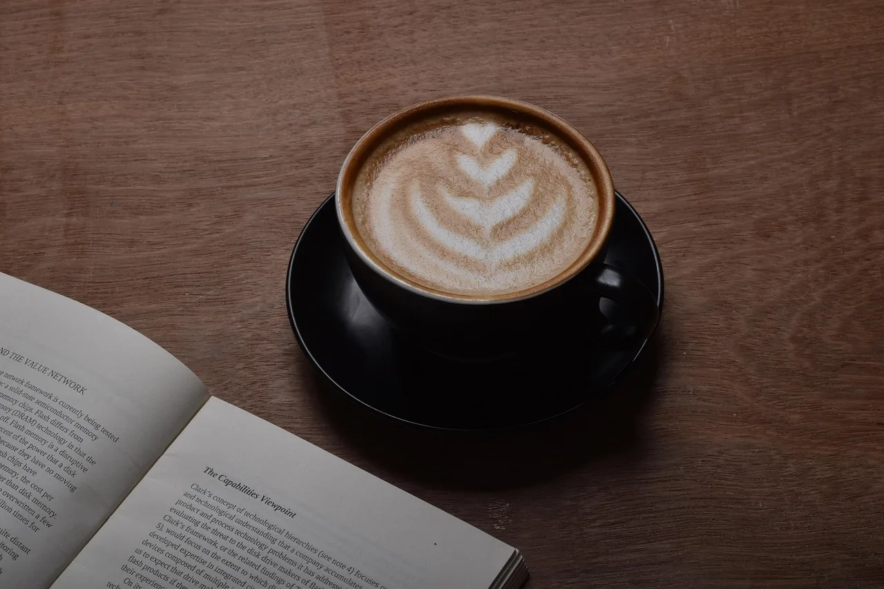 Coffee cup with latte art next to an open book on a wooden surface.