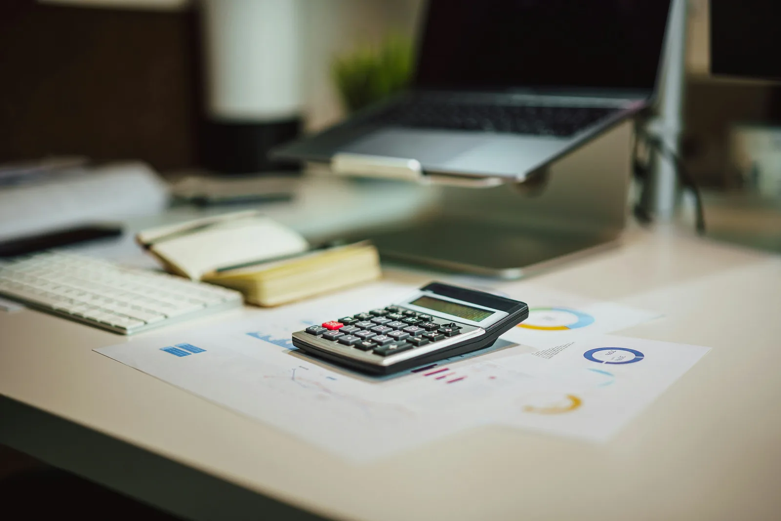 Calculator resting on printed data sheets and charts on a white desk next to a laptop.