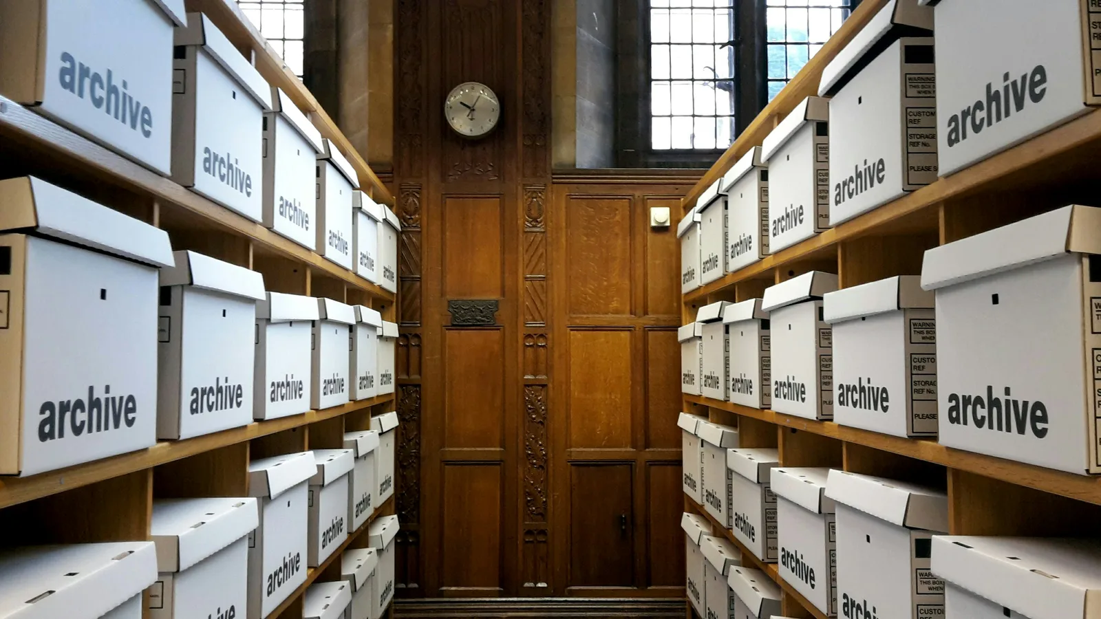Rows of white archive boxes stacked on wooden shelves in a room with a clock on the wall.